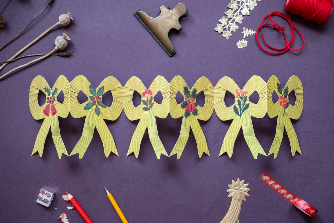 A row of yellow paper bow ornaments with colorful floral details, arranged on a purple background with various craft supplies like scissors, red thread, and wooden beads. Designed, created and retailed in the UK by Hadley Paper Goods. 
