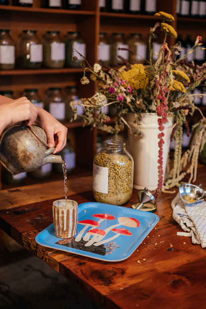 A hand pours liquid from a rustic metal pitcher into a small striped glass cup placed on a blue tray with a red-and-white mushroom print, set on a wooden table with dried flowers, jars, and shelves in the background.