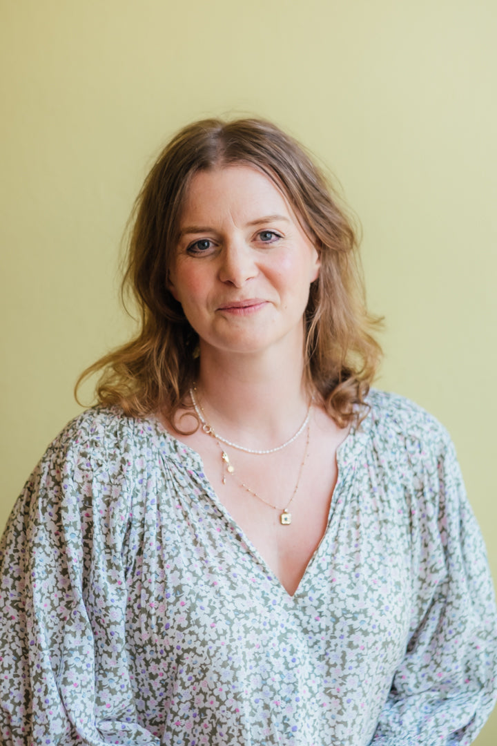 A woman with shoulder-length light brown hair wearing a light blue and white floral print blouse with layered gold necklaces, set against a solid pale yellow background.