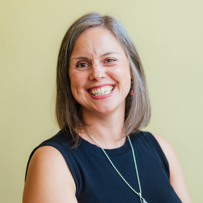 A smiling woman with shoulder-length gray-brown hair wearing a sleeveless black top and a silver pendant necklace, photographed against a plain light yellow-green background.