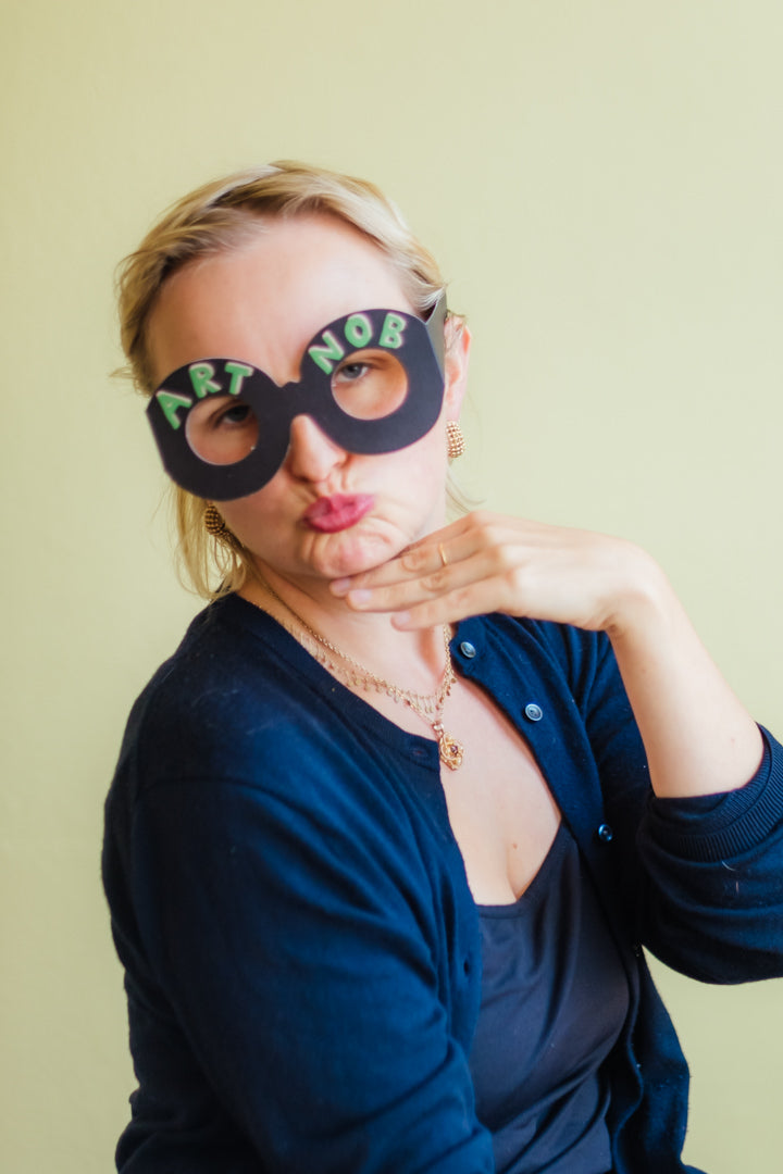 A woman wearing oversized black novelty glasses with the words 'ART NOB' in green and white lettering, posing with her hand near her chin against a pale yellow-green background.