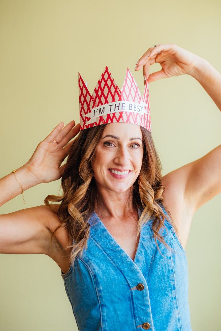A smiling woman with wavy brown hair wears a red and white patterned paper crown that says 'I'M THE BEST' against a solid pale yellow background.