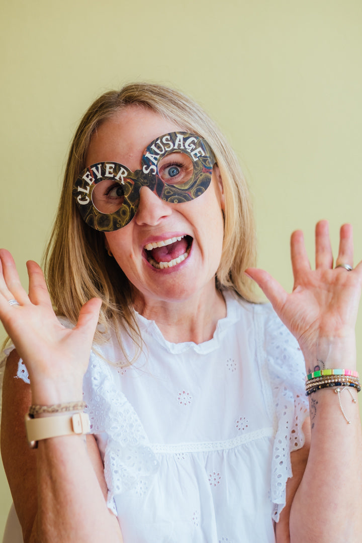 A woman with blonde hair wearing a white eyelet top and colorful bracelets, smiling widely while wearing novelty skull-shaped glasses that read 'CLEVER' on one side and 'SAUSAGE' on the other, against a pale yellow background.