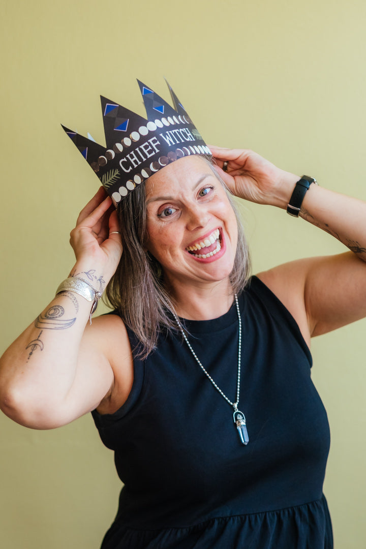 A woman smiling and adjusting a black paper crown with white text that reads 'CHIEF WITCH' against a solid olive-green background.