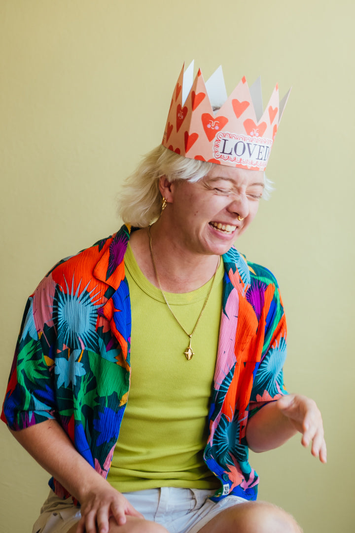 A person wearing a colorful tropical print shirt, lime green t-shirt, and a paper crown printed with red hearts and the word 'LOVE' on the front, set against a solid pale yellow background.