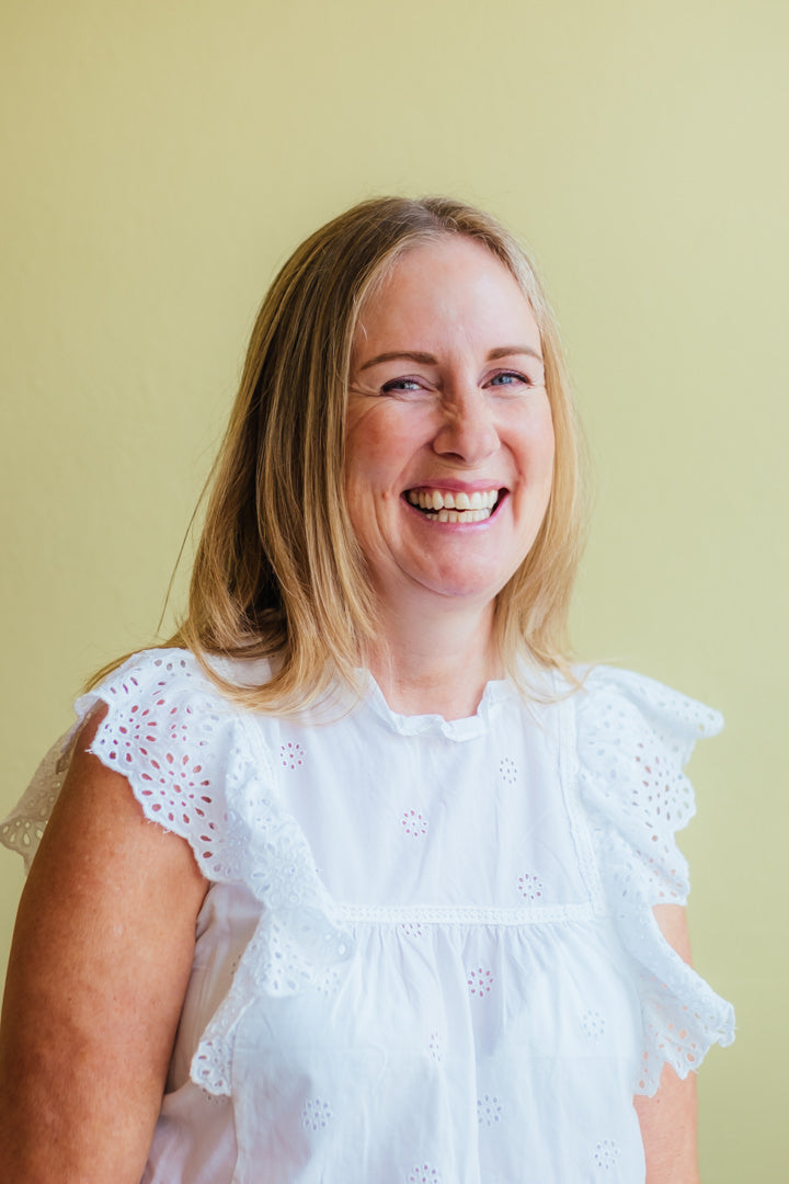 A smiling woman with blonde hair wearing a white eyelet blouse, photographed against a solid pale yellow-green background.